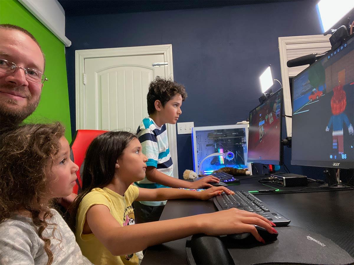 Photo of Slawth Dad (Mission Mike) playing videogames with Michael (Right), Maia (Middle) and Melody (Left).
                   They are sitting at a computer desk with a green screen behind them.